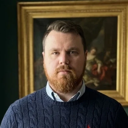 Shane Lint, founder and editor of The Oakmoss & Ivy, in a navy cable-knit sweater before a gilt-framed painting